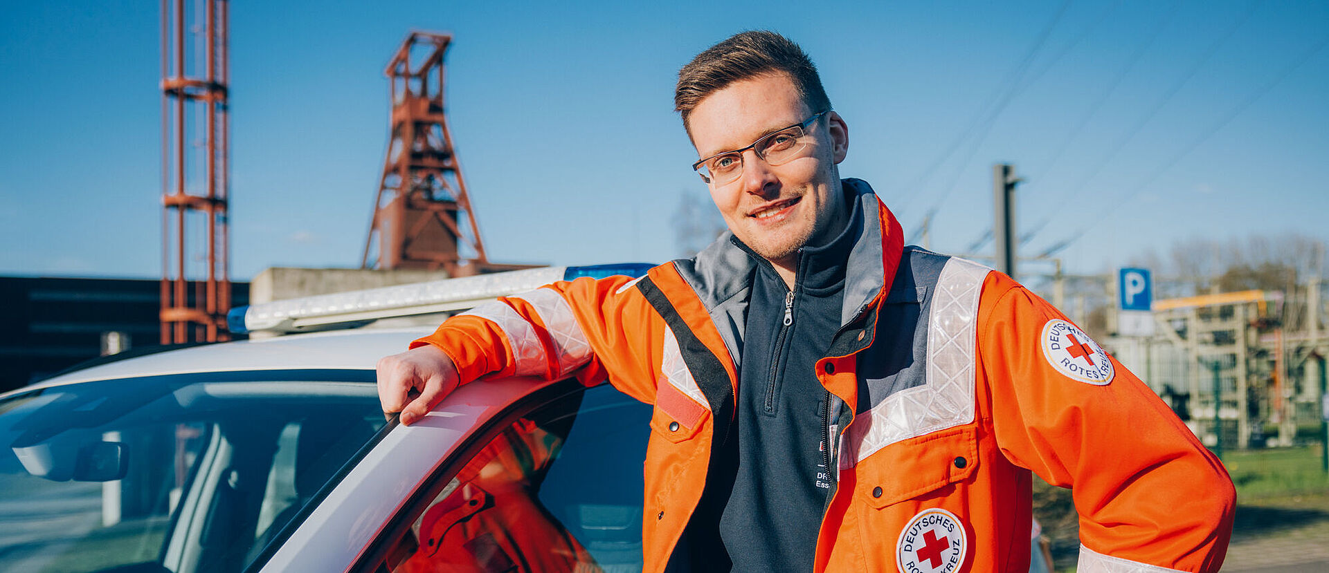 DRK Essen Ein lächelnder junger Mitarbeiter des DRK Essen lehnt in Einsatzkleidung an einem Auto des DRK. Im Hintergrund ist die Zeche Zollverein erkennbar.